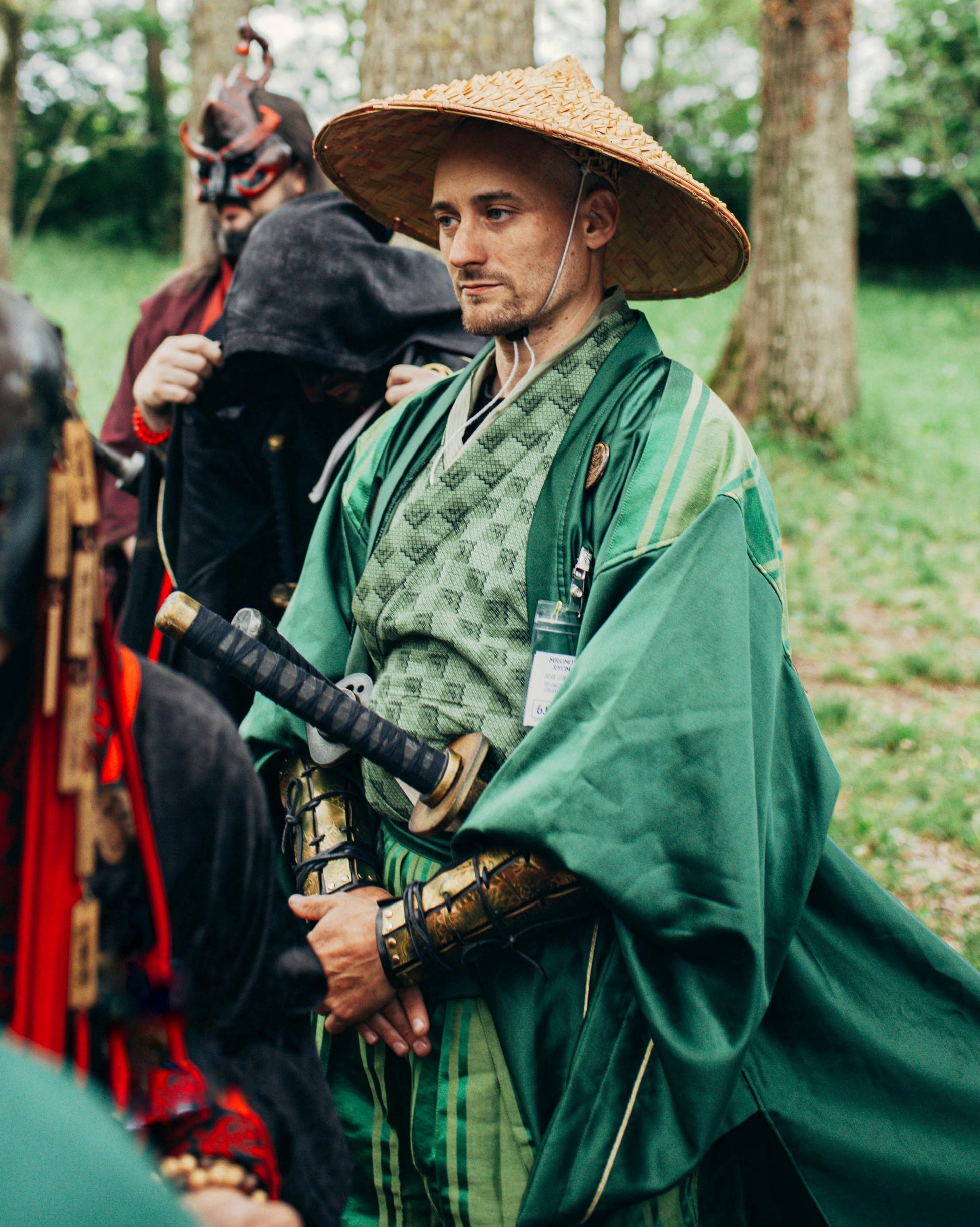 Portrait of LARP samurai in green outfit and wide hat, hands joined, wearing dragon bracers and traditional robe. Costume set ideal for samurai cosplay or fantasy warriors. Bracers crafted in golden engraved leather with vegetable tanning. Long tail SEO: samurai bracers cosplay, larp leather bracers, fantasy warrior costume accessories. Handmade for immersive roleplay settings.