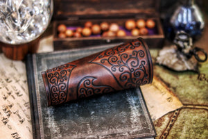 A pair of brown leather bracers with intricate embossed patterns, displayed on a table with a crystal ball and other fantasy-themed items in the background. Shadowbinder bracer for mages & wizards, sorcerer vambarce for larp costume and wizardry cosplay leather witchcraft handmade