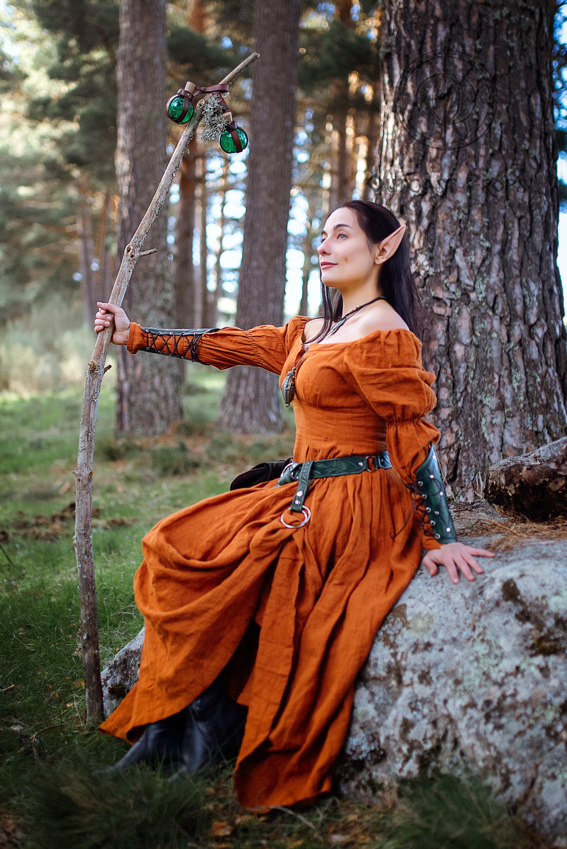 A woman wearing a long, rust-colored dress with a green belt and a brown leather strap with a metal ring. She is holding a wooden staff with green glass orbs attached to it. The woman is sitting on a large rock in a forest setting.