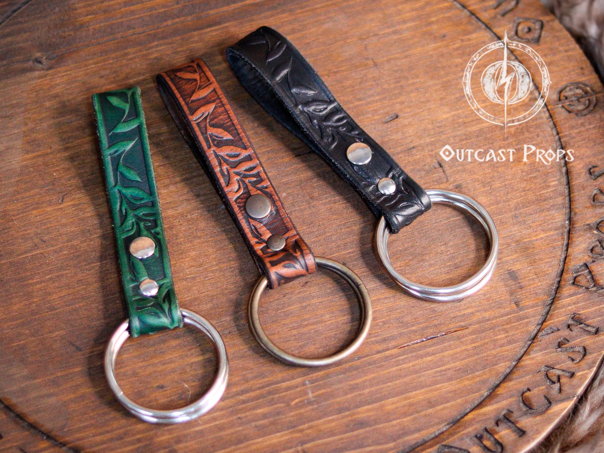 Three Skirt Hike with metal rings. The skirt hikes  are brown, green, and black with a floral design. The skirt hikes are displayed on a wooden surface with the text 'Outcast Props'. Medieval Floral Leather Skirt Hike Ring LARP Ren Faire Cosplay Accessory for Fantasy Outfits Skirt Hiker Renaissance Fair Costume Accessory