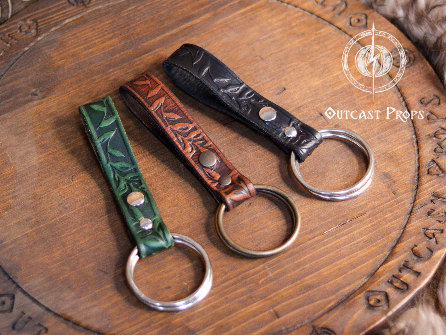 Three Skirt Hikes with metal rings. The skirt hikes  are brown, green, and black with a floral design. The skirt hikes are displayed on a wooden surface with the text 'Outcast Props'. Medieval Floral Leather Skirt Hike Ring LARP Ren Faire Cosplay Accessory for Fantasy Outfits Skirt Hiker Renaissance Fair Costume Accessory