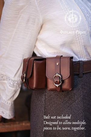 Front view of a medieval leather belt pouch worn with historical inspired clothing, showing the brass buckle closure and smooth brown leather finish. Handmade using traditional techniques and aged by hand, this pouch can be used as a deck bag for Magic The Gathering, Pokémon TCG, and Arkham Horror card games. Compact yet sturdy, it is ideal for LARP characters, Renaissance fair outfits, steampunk costumes, or daily carry. Designed to fit standard belts and to be combined with multiple similar leather pouche