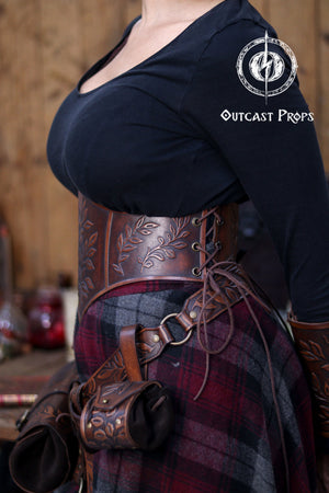 Close side view of the floral leather underbust corset belt highlighting the botanical embossing and warm aged brown tones. The lacing on the side shows the adjustable nature of the corset, making it suitable for LARP costumes, elven outfits, witchcore fashion and renaissance faire clothing. Matching accessories and fantasy skirt complete the handcrafted look. Outcast Props logo appears in the corner.