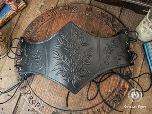 Front view of a black floral embossed leather underbust corset crafted from premium vegetable tanned leather, ideal for elves, witches, herbalists and noble characters at Renaissance Faire, renfaire or LARP events. The detailed leaf and flower pattern stands out against the dark finish, with side lacing for an adjustable fit. The corset lies flat on a wooden table surrounded by props, showing its elegant medieval fantasy design ready to pair with belts and accessories.