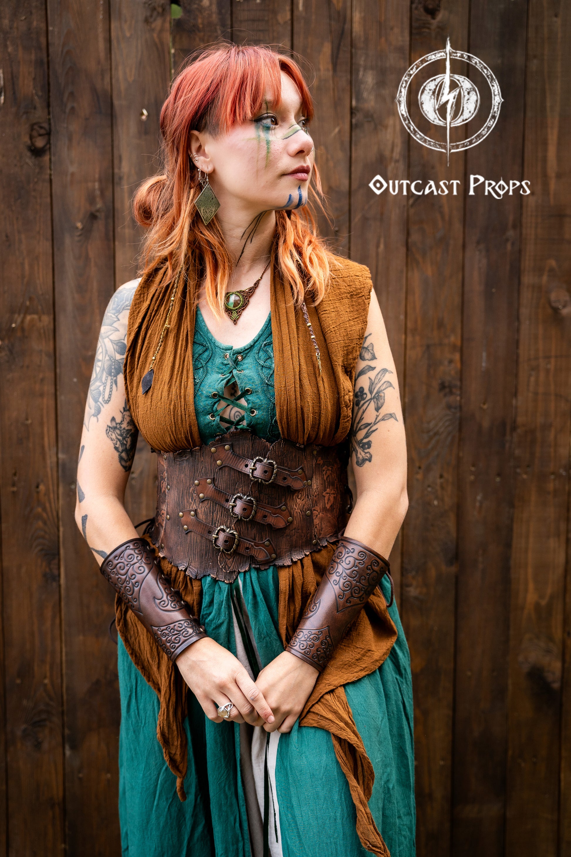 Woman wearing a handcrafted leather corset belt with carved wood texture and embossed ivy motifs, paired with embossed bracers. She has red hair, green and brown layered dress, and fantasy makeup on her face. The corset features three decorative buckles and side lacing. Ideal for elven costume, renaissance fair clothing, herbalist cosplay outfit, and LARP druid armor. The full costume is styled with nature-inspired elements and artisanal detailing.