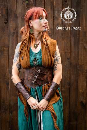 Woman wearing a handcrafted leather corset belt with carved wood texture and embossed ivy motifs, paired with embossed bracers. She has red hair, green and brown layered dress, and fantasy makeup on her face. The corset features three decorative buckles and side lacing. Ideal for elven costume, renaissance fair clothing, herbalist cosplay outfit, and LARP druid armor. The full costume is styled with nature-inspired elements and artisanal detailing.