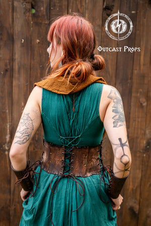 Back view of a green dress and brown underbust corset belt made of leather with woodgrain texture and floral engraving. The corset is tied with adjustable leather laces at the back and sides. The model’s red hair is in a low bun, and both arms show large tattoos. The garment combination suits elf costume, renaissance druid corset, and herbalist cosplay. The image highlights how the corset belt fits and closes, showing its shape and back fastening method.
