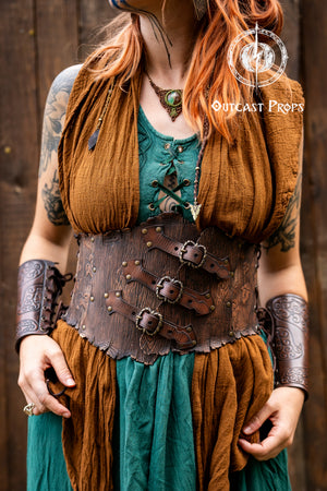 Person in a fantasy costume with 'Outcast Props' logo in the corner. Person wearing a detailed leather corset with decorative elements, branded 'Outcast Props'. Underbust Wood Leather Corset. Herbalist corset. Druid corset. Elven Corset. Ivy Corset.