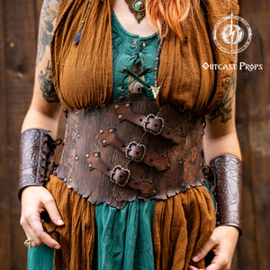 Close-up of a woodgrain leather underbust corset belt with three horizontal buckled straps, floral embossing, and riveted accents. The model wears a green lace-up top under a brown draped shawl. Visible details include a green pendant necklace, large metal earrings, and stylized face paint. A detailed view of the textures, leather tooling, and layered costume elements, perfect for fantasy corset belt, druid outfit, elf LARP clothing, and steampunk leather armor.