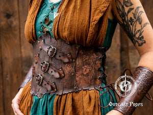 Angled close-up of a fantasy corset belt with rich brown leather, embossed bark texture, and three front buckles with rounded straps. The model wears a green medieval top with chest lacing and a layered brown shawl. Visible tattoos on her arm and a green gemstone pendant enhance the herbalist or elven LARP style. Ideal for wood leather corset belt, renaissance costume, fantasy cosplay accessory. The focus is on texture, construction, and integration into the outfit.