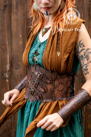 Side view of a woman in a green and brown medieval-style dress, wearing a wide leather underbust corset belt with carved floral patterns and three front buckles. The model also wears embossed leather bracers with swirling motifs. The outfit is styled for a fantasy character such as a woodland druid, ranger, or herbalist. Ideal for renaissance fair clothing, LARP accessories, and leather corset belt cosplay. The photo shows the belt’s shape, textures, and layered fabrics.