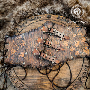 Flat lay of the underbust leather corset belt on a round engraved wooden board with Outcast Props branding. The corset shows detailed embossed ivy vines and wood grain texture. Three adjustable buckles and brass rivets on the front, and side lacing holes visible. Ideal for elf costume belt, nature druid outfit, medieval fantasy corset, or LARP herbalist gear. Handmade from vegetable-tanned leather and aged by hand.