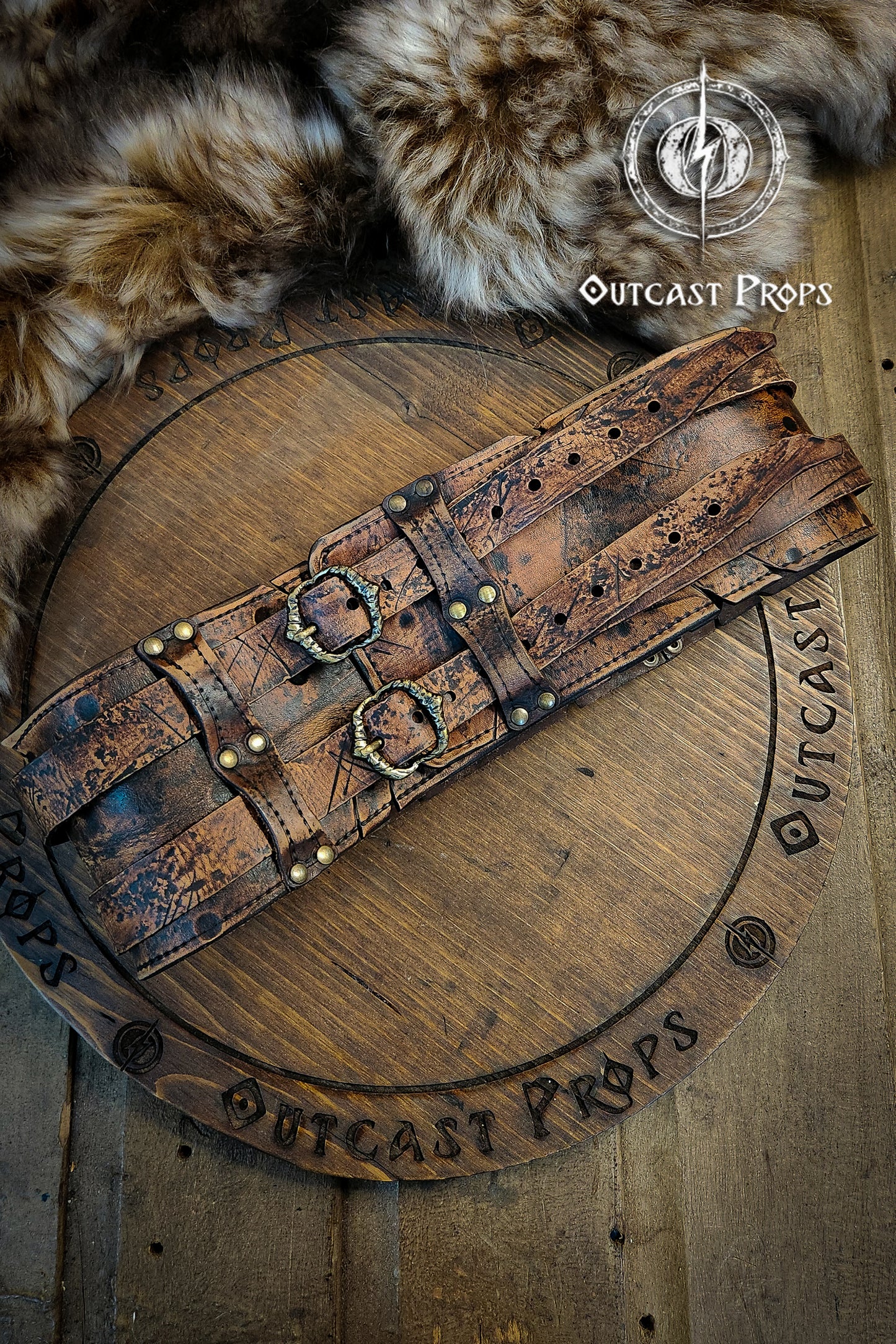 Brown leather belt with metal buckles on a wooden surface, branded 'Outcast Props'. Veteran Leather Belt premium vegetable-tanned, worn by raiders and mercenaries.