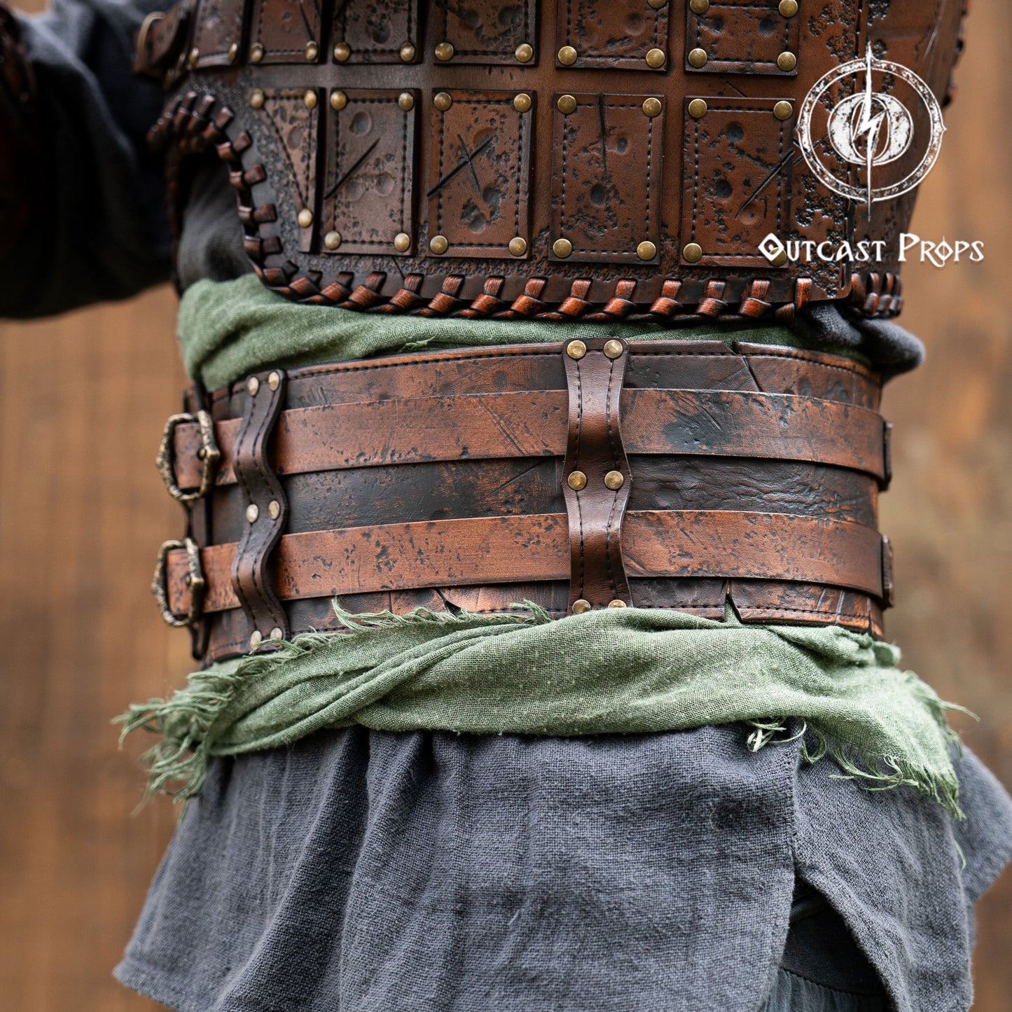 Close-up of a person wearing detailed leather armor with 'Outcast Props' branding. Close-up of a detailed costume with leather straps and a green cloth, branded 'Outcast Props'. Veteran Leather Belt premium vegetable-tanned, worn by raiders and mercenaries.
