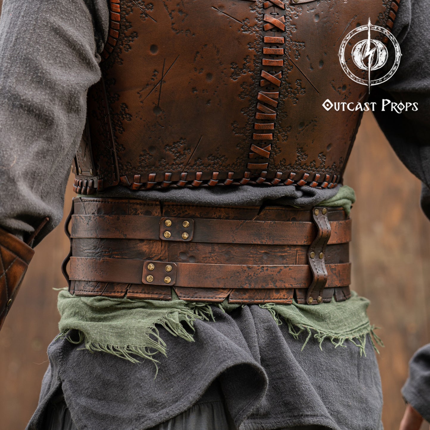 Close-up of a detailed leather vest with metal fastenings, worn by a person, on a blurred background. Close-up of a detailed costume with leather straps and a green cloth, branded 'Outcast Props'. Veteran Leather Belt premium vegetable-tanned, worn by raiders and mercenaries.