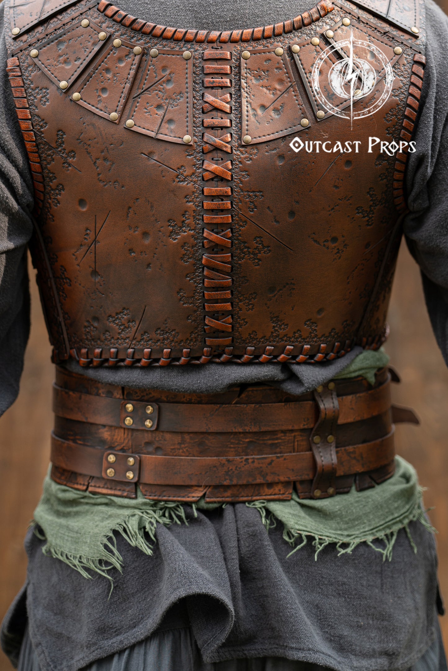 Brown leather vest with intricate details and 'Outcast Props' branding on a blurred background. Close-up of a detailed costume with leather straps and a green cloth, branded 'Outcast Props'. Veteran Leather Belt premium vegetable-tanned, worn by raiders and mercenaries.