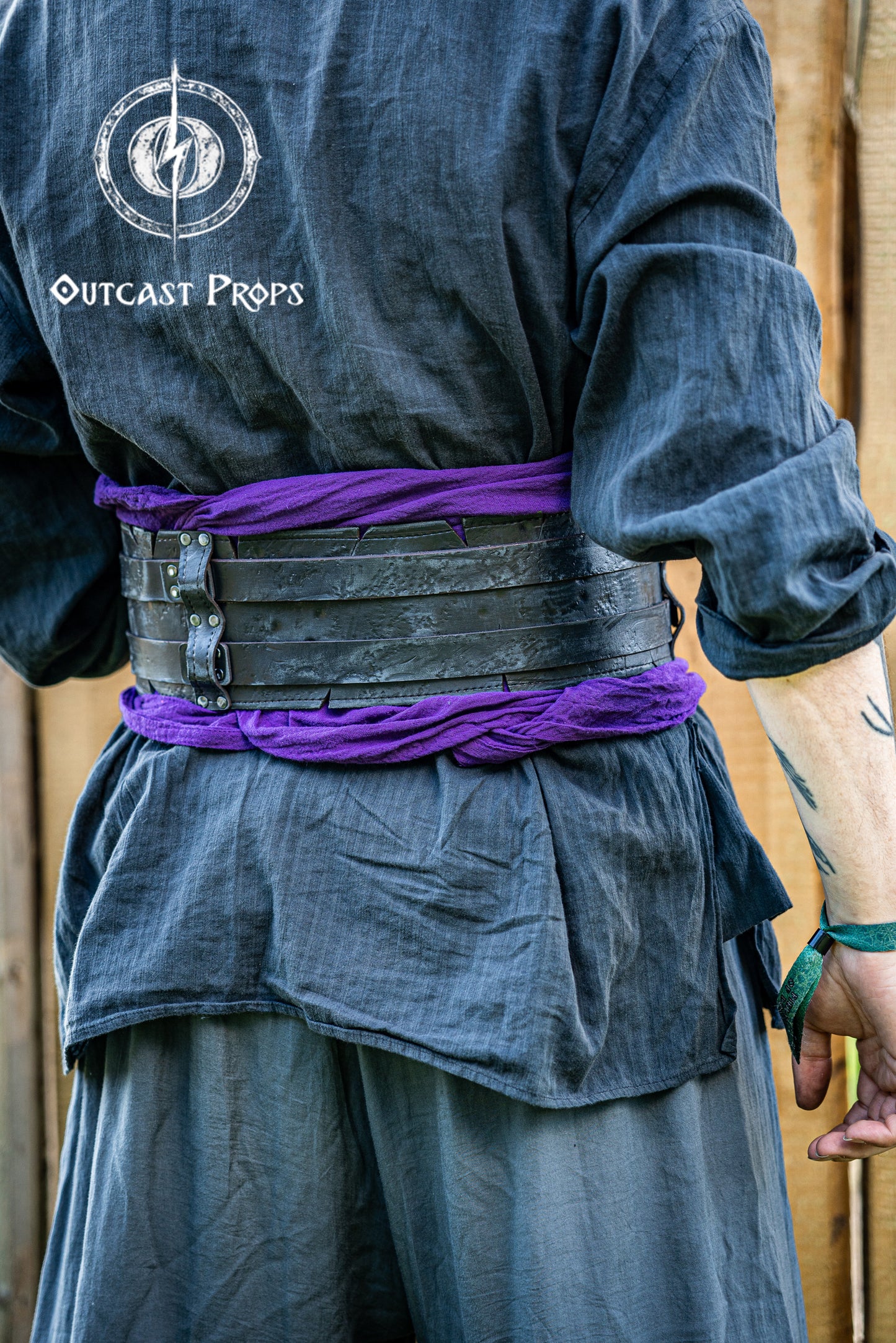 Person wearing a dark outfit with a purple sash, standing against a wooden background with 'Outcast Props' branding. Person wearing a dark outfit with a purple sash and black leatrher broadbelt with Outcast Props logo in the corner. Veteran Leather Belt premium vegetable-tanned, worn by raiders and mercenaries.