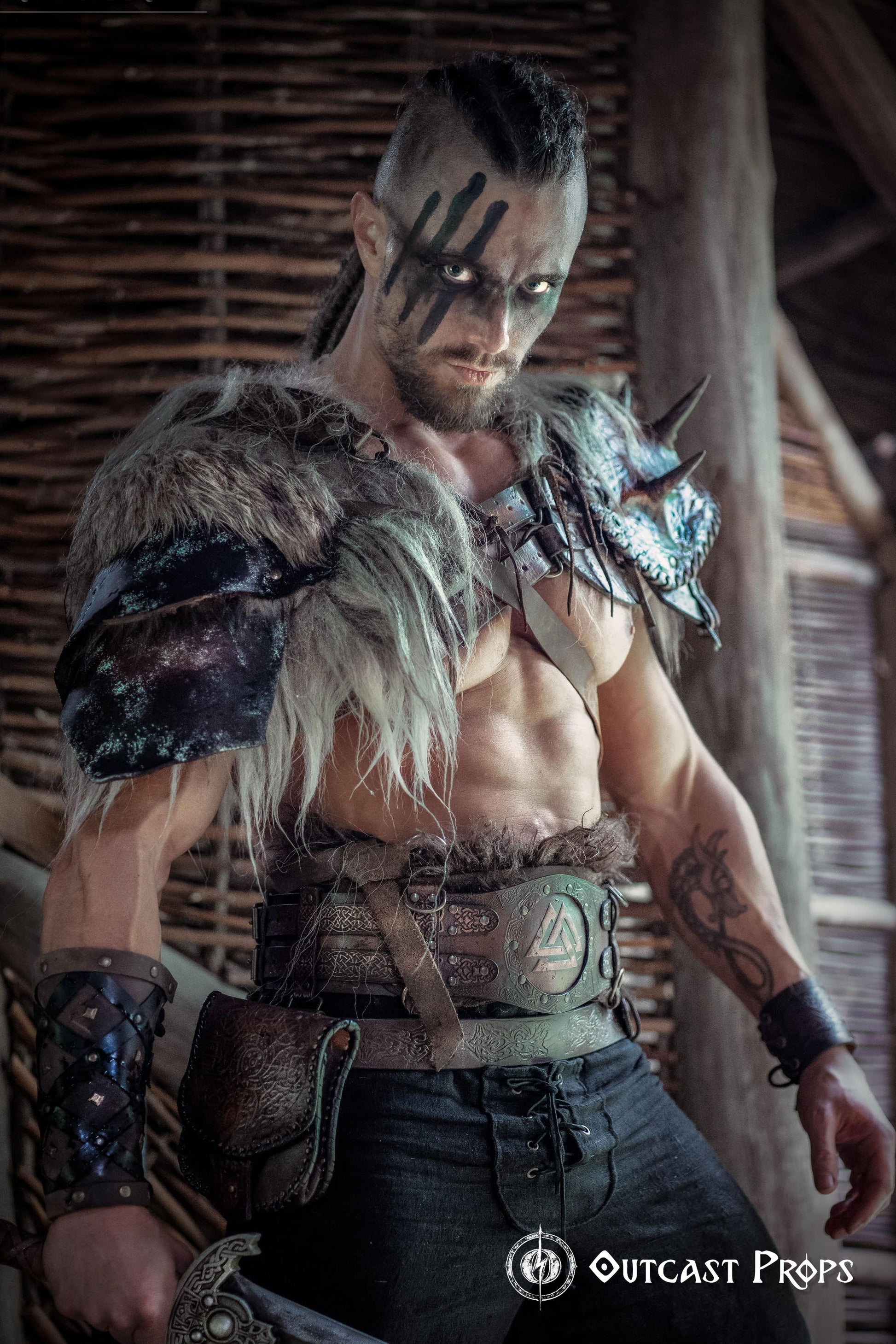Barbarian cosplay outfit worn by a male model showing the viking leather armor belt with Valknut design. The wide belt cinches the waist and supports pouches and gear, matching the celtic fantasy belt theme. Ideal for LARP, fantasy warrior look, or renfaire costume. The photo captures the rugged, immersive style in a dark wooden hut.