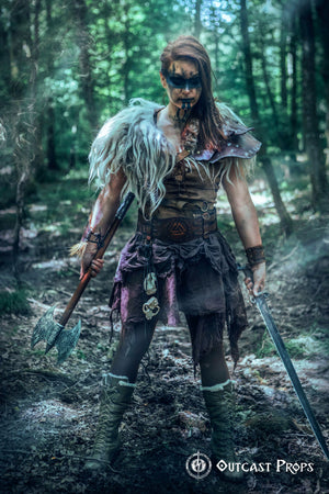 Barbarian cosplay outfit worn by a woman model showing the viking leather armor belt with Valknut design. The wide belt cinches the waist and supports pouches and gear, matching the celtic fantasy belt theme. Ideal for LARP, fantasy warrior look, or renfaire costume. The photo captures the rugged, immersive style in a dark wooden hut.