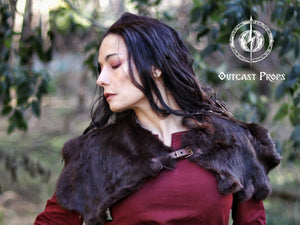 Female model in a red medieval tunic wearing a dark fur Viking mantle in a forest setting. The cape features a brown leather clasp and realistic rugged texture, perfect for Celtic warrior cosplay, tribal characters, or LARP barbarian costumes. A handmade accessory for fantasy fairs, Viking roleplay, or historical reenactment. Styled for immersion and warmth in outdoor adventures.