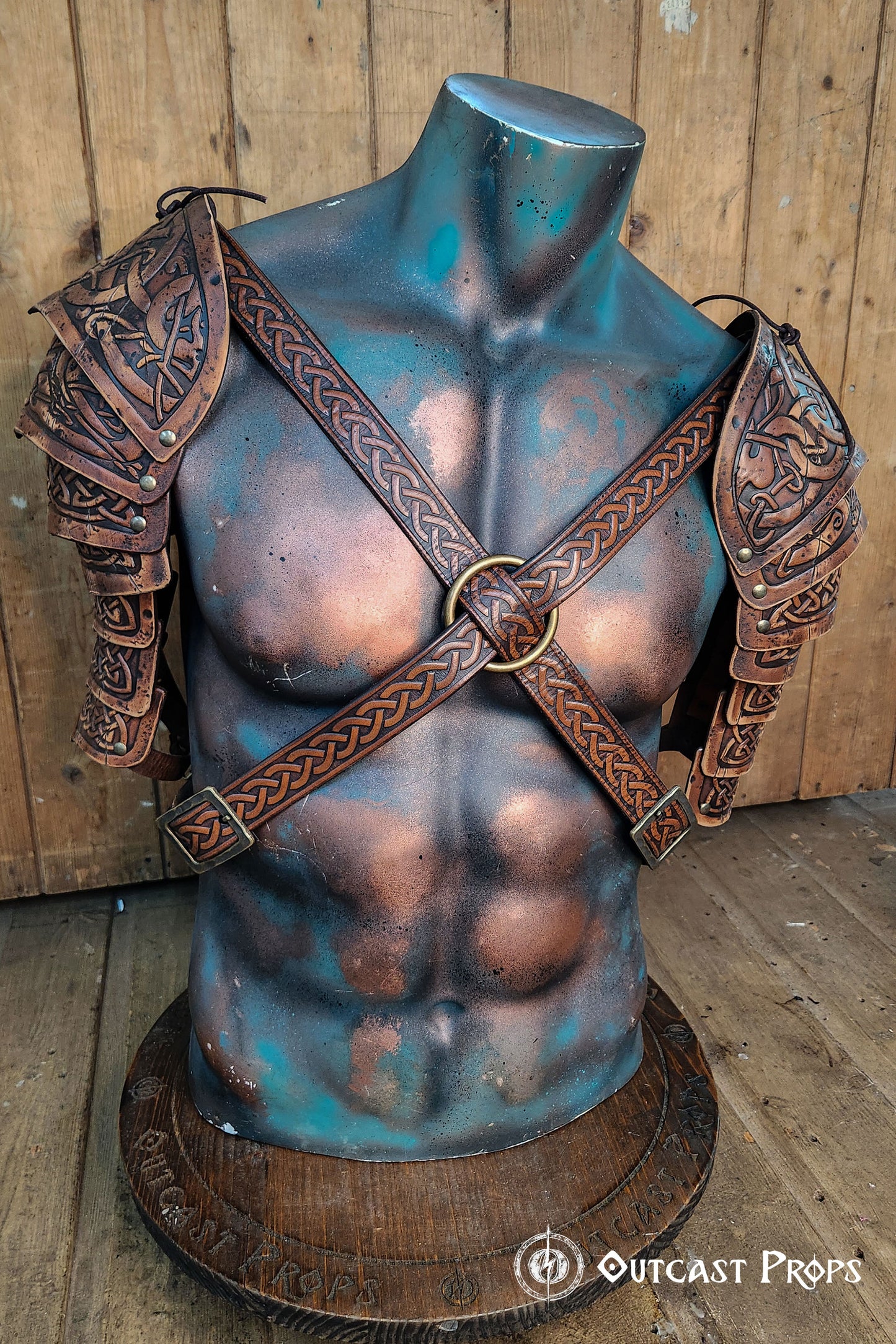 Detailed close-up of Viking leather shoulder armor Sedrik for LARP cosplay, showing Celtic knot engravings on layered brown leather pauldrons with aged finish. Armor displayed on a sculpted mannequin torso, crossed leather straps with embossed knots and metal ring at chest, rustic wooden backdrop. Ideal for Viking warrior costume, fantasy LARP outfit, Celtic cosplay accessories, premium handcrafted leather armor for historical or fantasy events.
