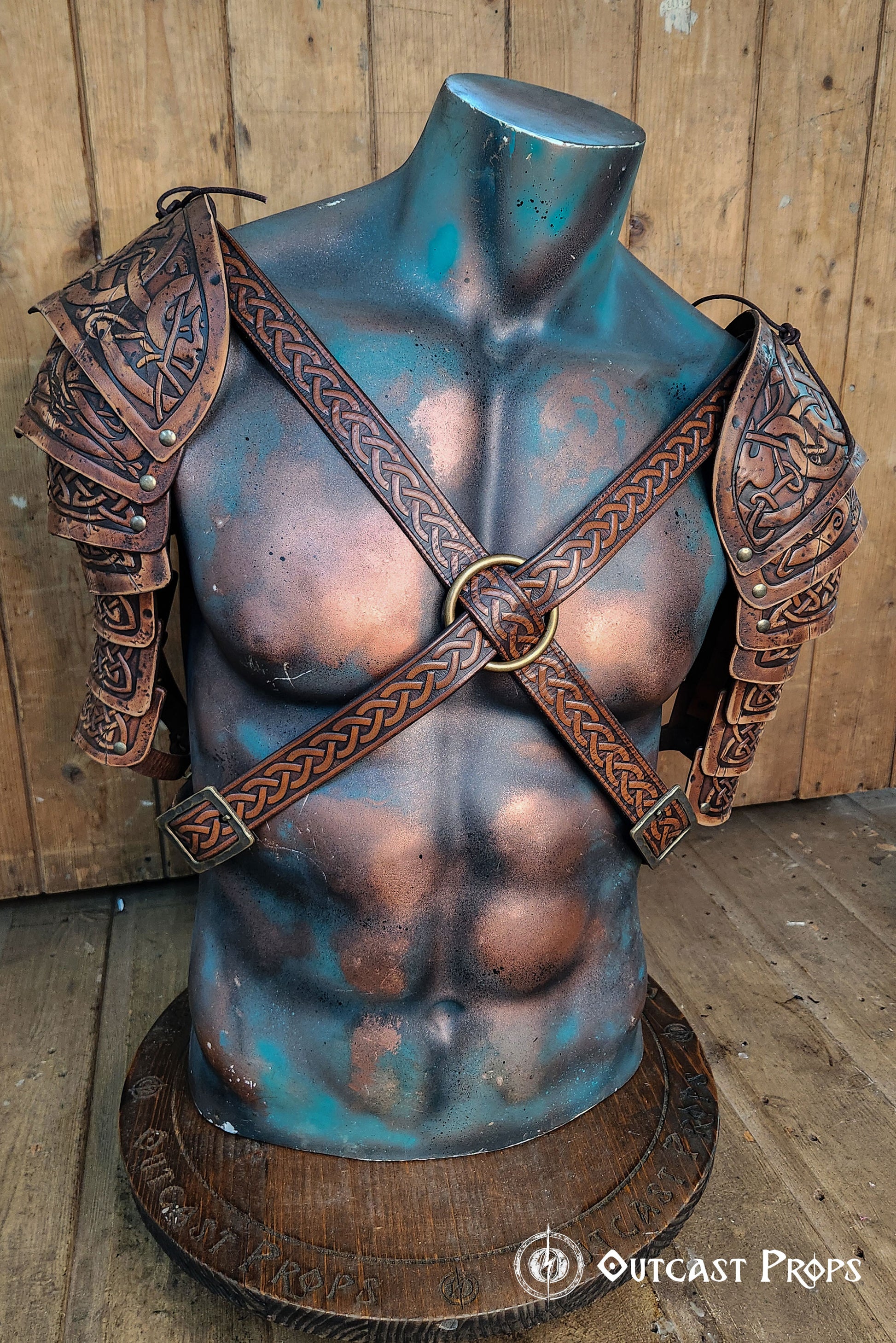 Detailed close-up of Viking leather shoulder armor Sedrik for LARP cosplay, showing Celtic knot engravings on layered brown leather pauldrons with aged finish. Armor displayed on a sculpted mannequin torso, crossed leather straps with embossed knots and metal ring at chest, rustic wooden backdrop. Ideal for Viking warrior costume, fantasy LARP outfit, Celtic cosplay accessories, premium handcrafted leather armor for historical or fantasy events.