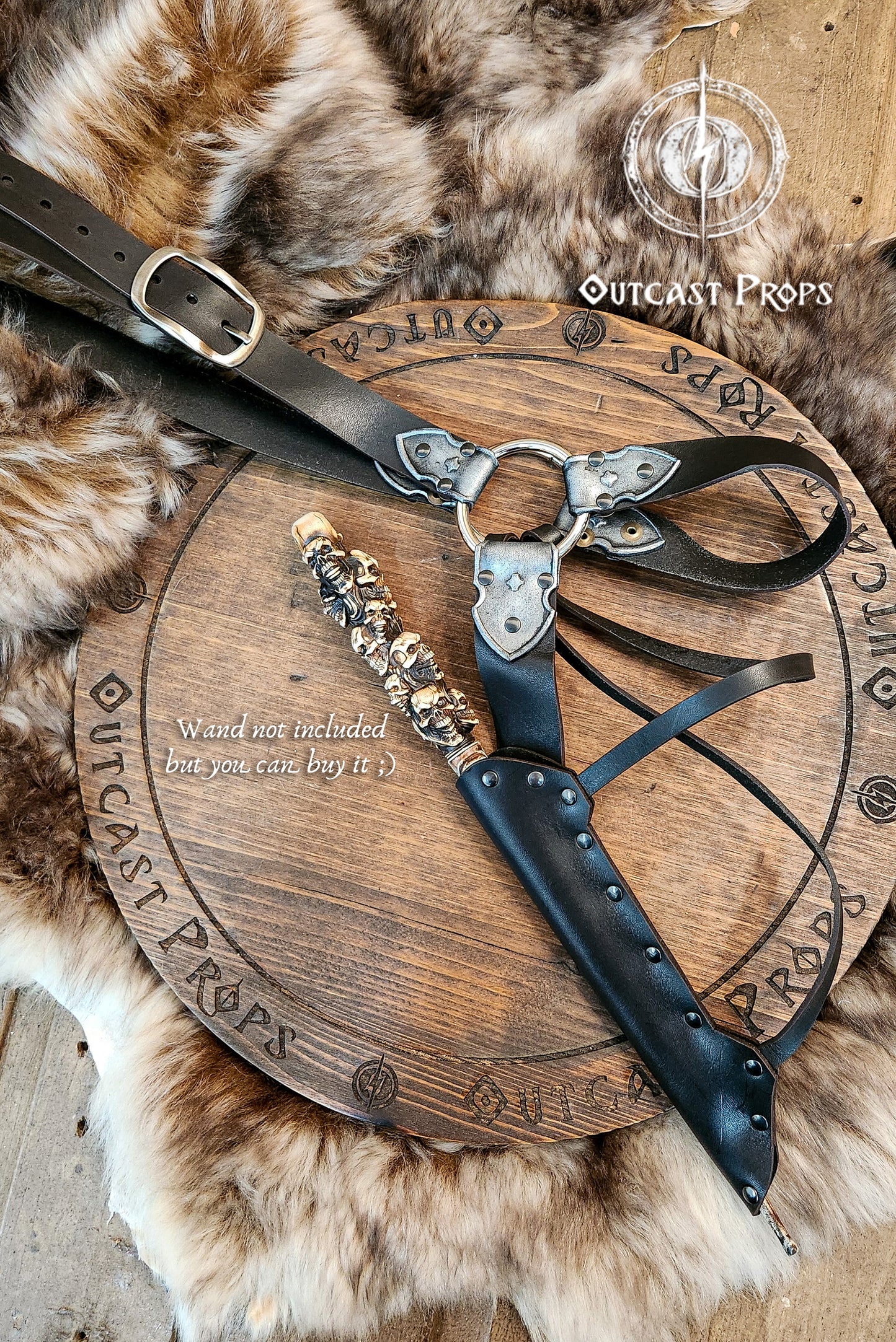 A black leather and silver wand holster with dual metal rings and riveted construction. Styled for Voldemort-inspired cosplay or LARP magic duels. Crafted for comfort and realism in wizard costumes, especially for Harry Potter fans recreating Death Eater aesthetics. Decorative sheath with leather strap on a wooden board, surrounded by fur, with 'Outcast Props' branding.