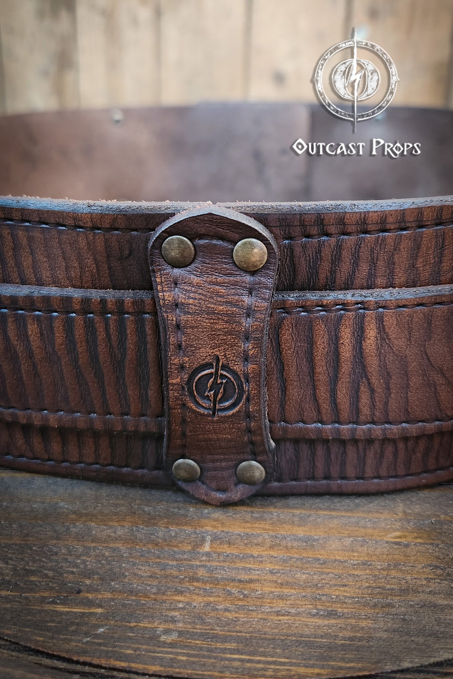 Close-up of a leather belt with metal fastenings on a wooden surface, branded 'Outcast Props'. Brown leather belt with brass buckle on a wooden surface, branded 'Outcast Props'. Medieval broadbelt for larp or cosplay. Elegant leather belt.