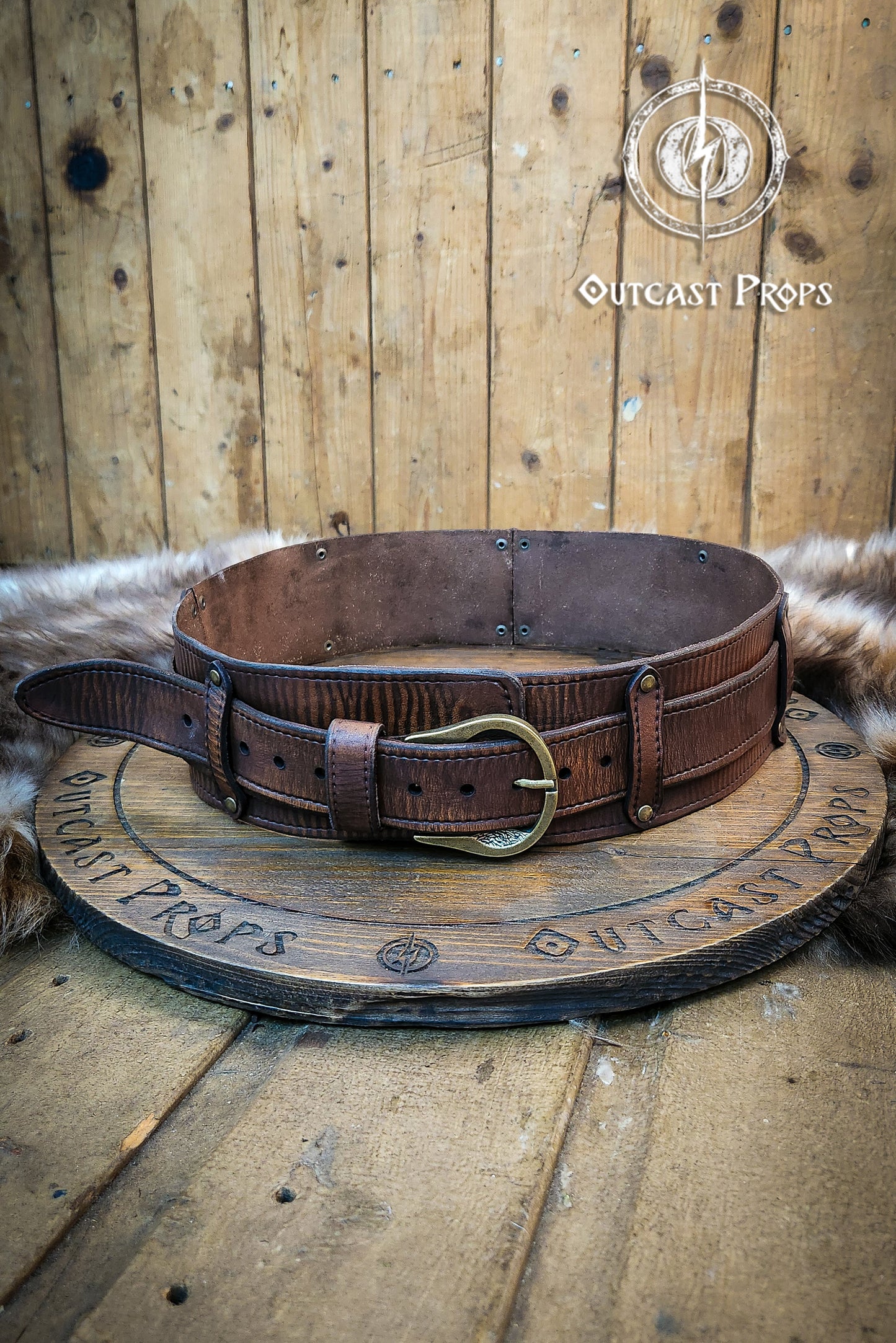 Brown leather belt with a gold buckle on a wooden surface, branded 'Outcast Props'.