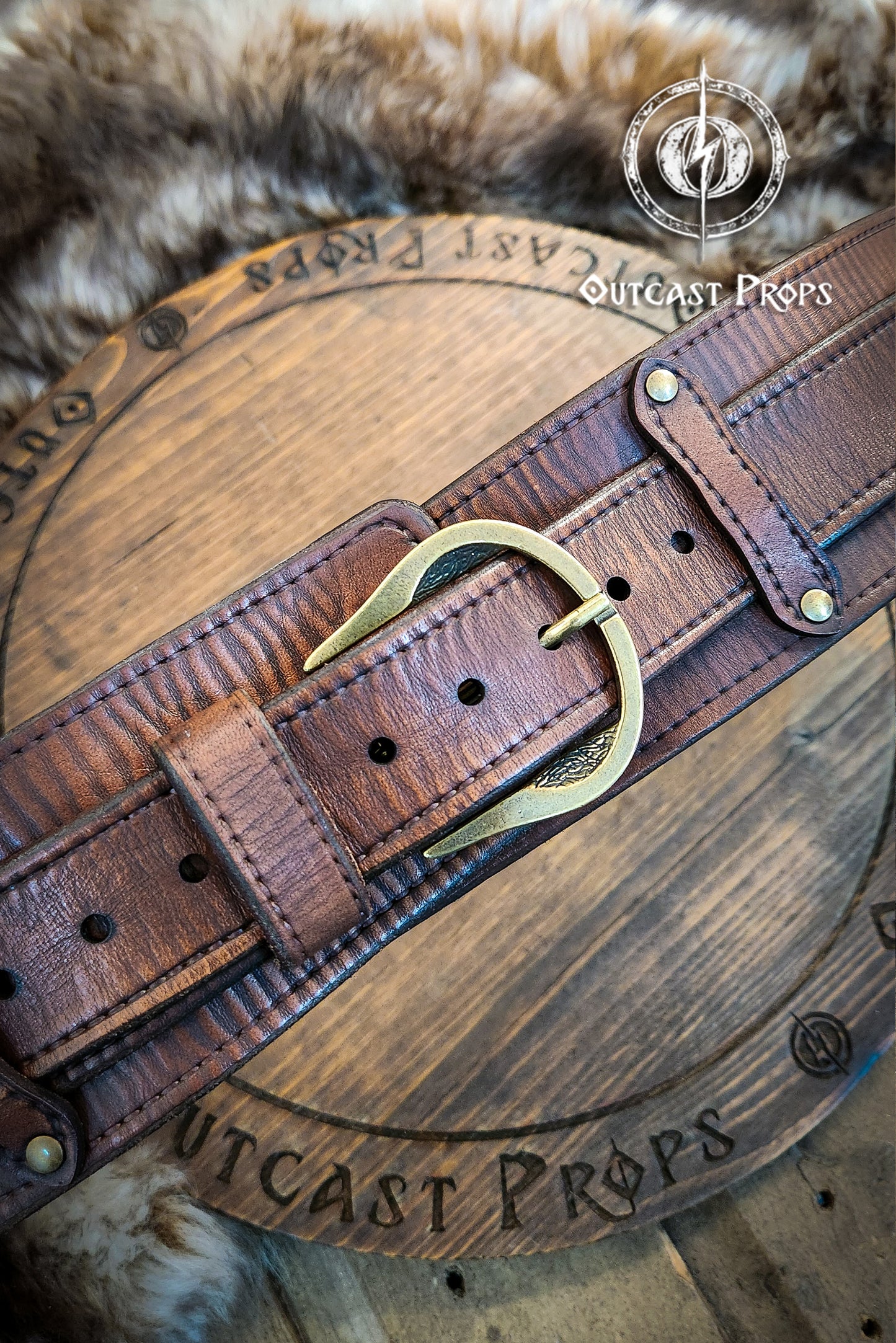 Brown leather belt with brass buckle on a wooden surface, featuring 'Outcast Props' branding. Brown leather belt with brass buckle on a wooden surface, branded 'Outcast Props'. Medieval broadbelt for larp or cosplay. Elegant leather belt.