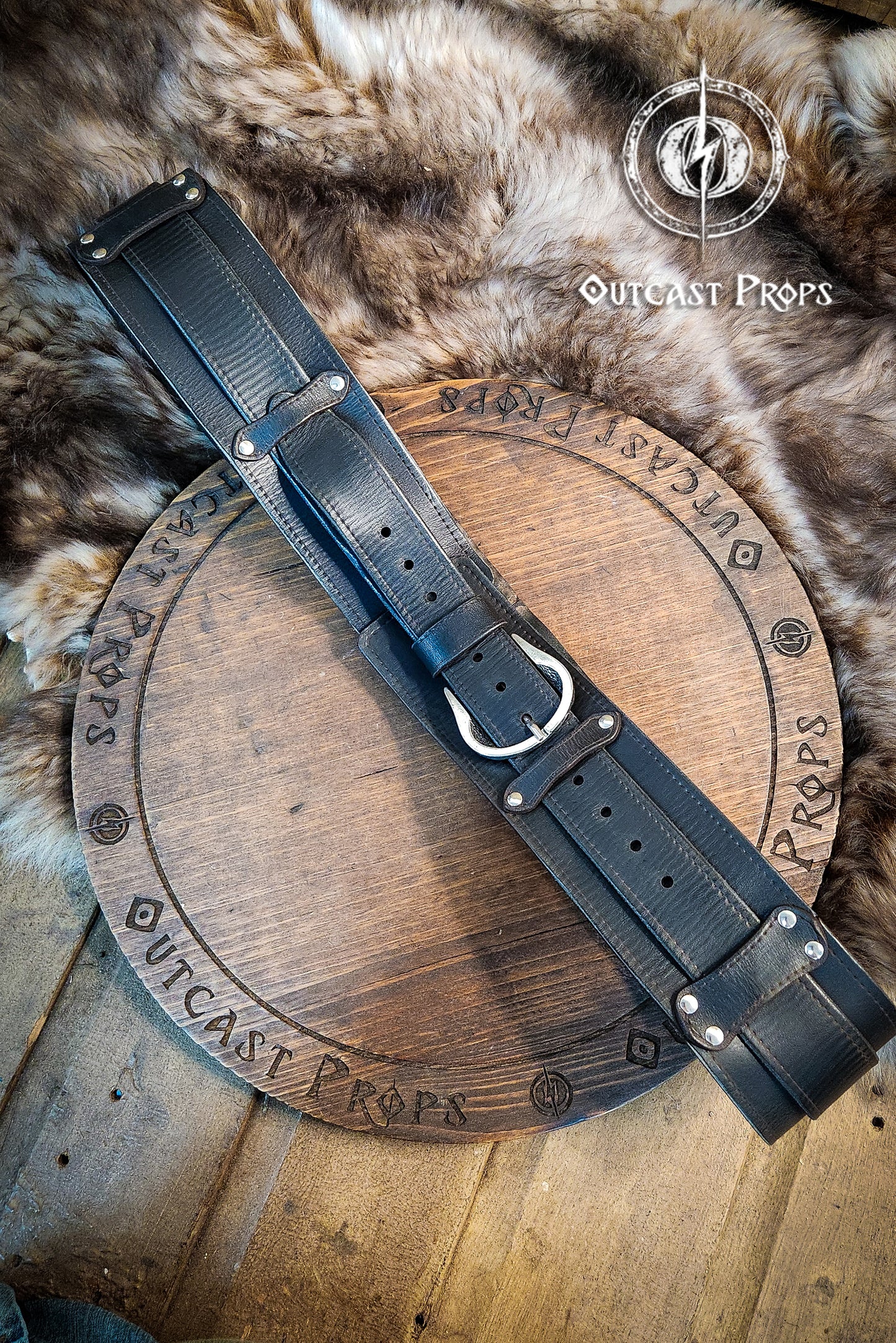 Black leather belt with metal buckle on a wooden disc, branded 'Outcast Props', on a fur background. Black leather belt with brass buckle on a wooden surface, branded 'Outcast Props'. Medieval broadbelt for larp or cosplay. Elegant leather belt.