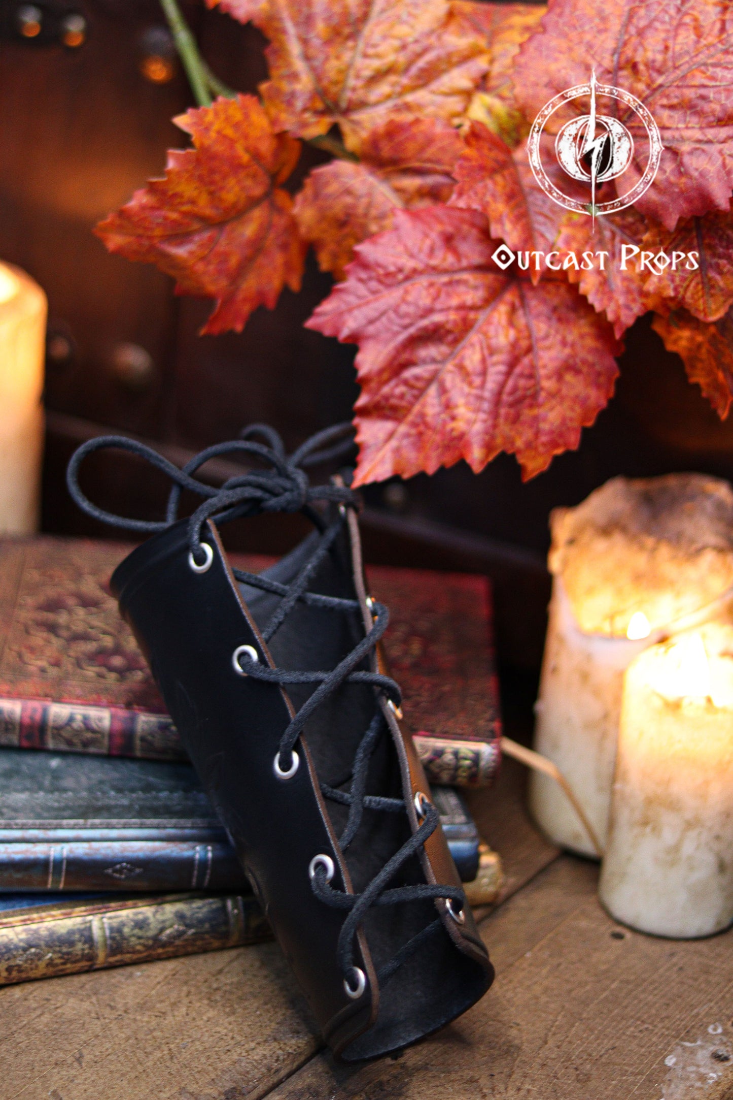 The back of the Wildflower leather bracer in black is shown with its laced fastening, resting on textured wooden boards with candles, autumn leaves and old books. This renaissance faire accessory is ideal for larp cosplay accessory outfits, elven herbalist witch roles and fantasy characters seeking elegant botanical leather gear. The scene emphasizes the handmade quality and natural curve of the bracer.