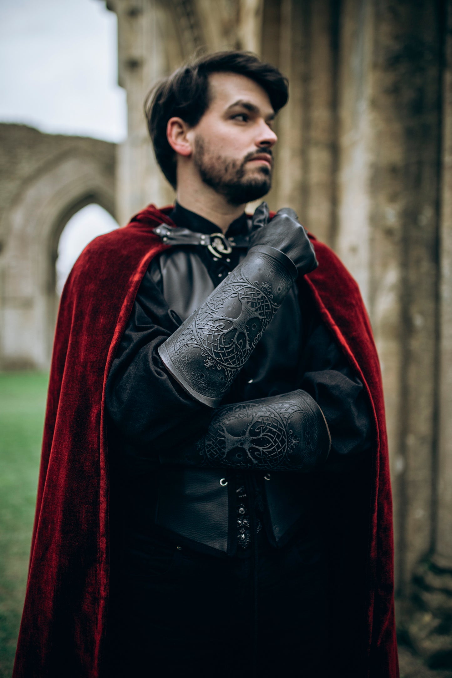 A fantasy LARP warrior wearing dark leather clothing and a red velvet cloak shows off black Yggdrasil leather bracers embossed with Norse world tree details. These long bracers, perfect for viking costume, leather armor, or medieval cosplay, offer both protection and character depth. Ideal for renaissance fairs, Geralt cosplay, or as female leather armor accessories. Model stands in front of a ruined stone archway, crossing arms with solem