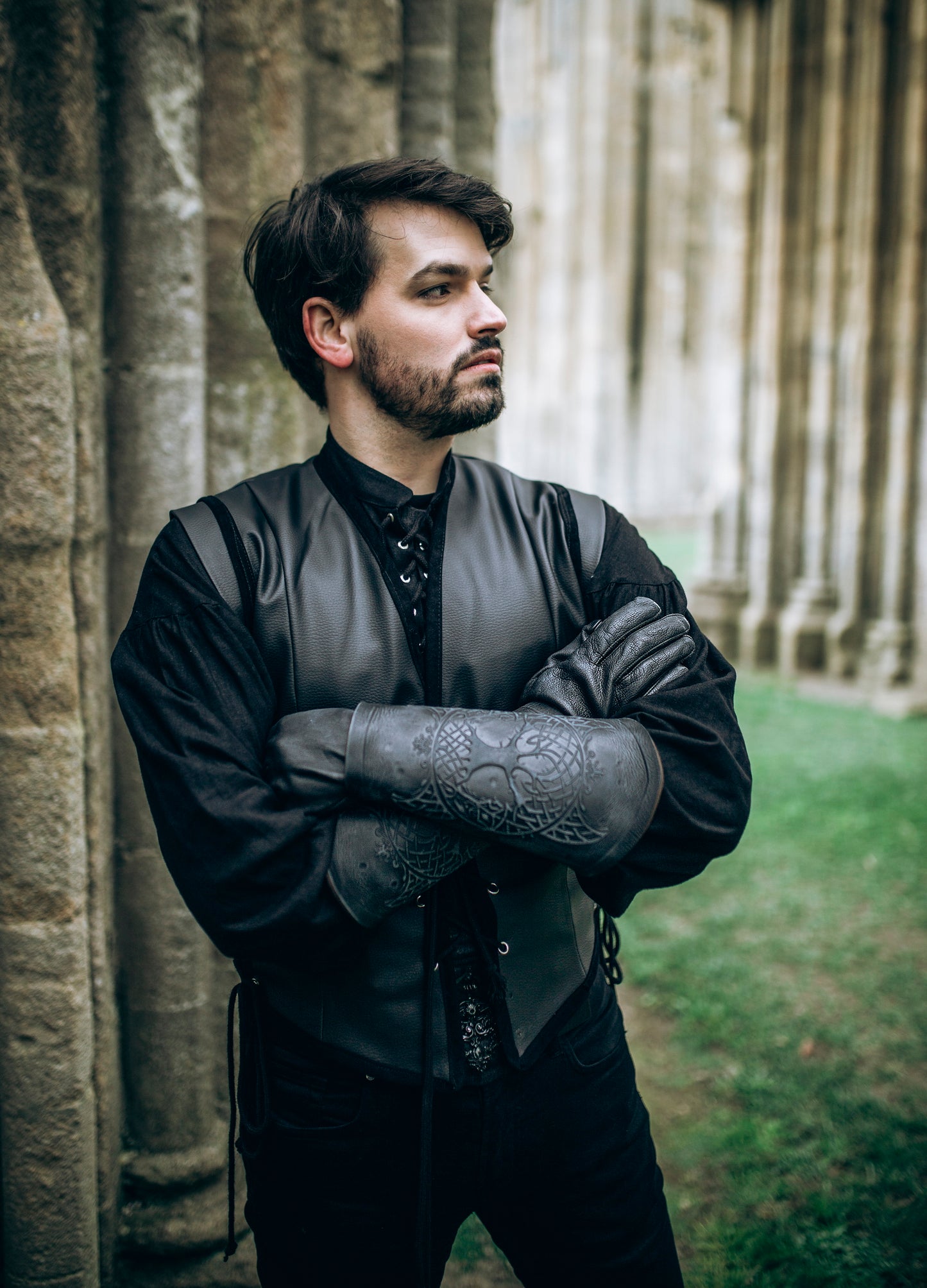 Man dressed in full black medieval outfit poses confidently with arms crossed, showcasing detailed black leather bracers embossed with Yggdrasil, the Norse tree of life. These vambraces are ideal for LARP armor, viking cosplay or as part of a Geralt witcher costume. The setting of ancient stone pillars and green grass adds an immersive backdrop for fantasy characters. Great for larping, female warrior armor, or medieval leather accessories.