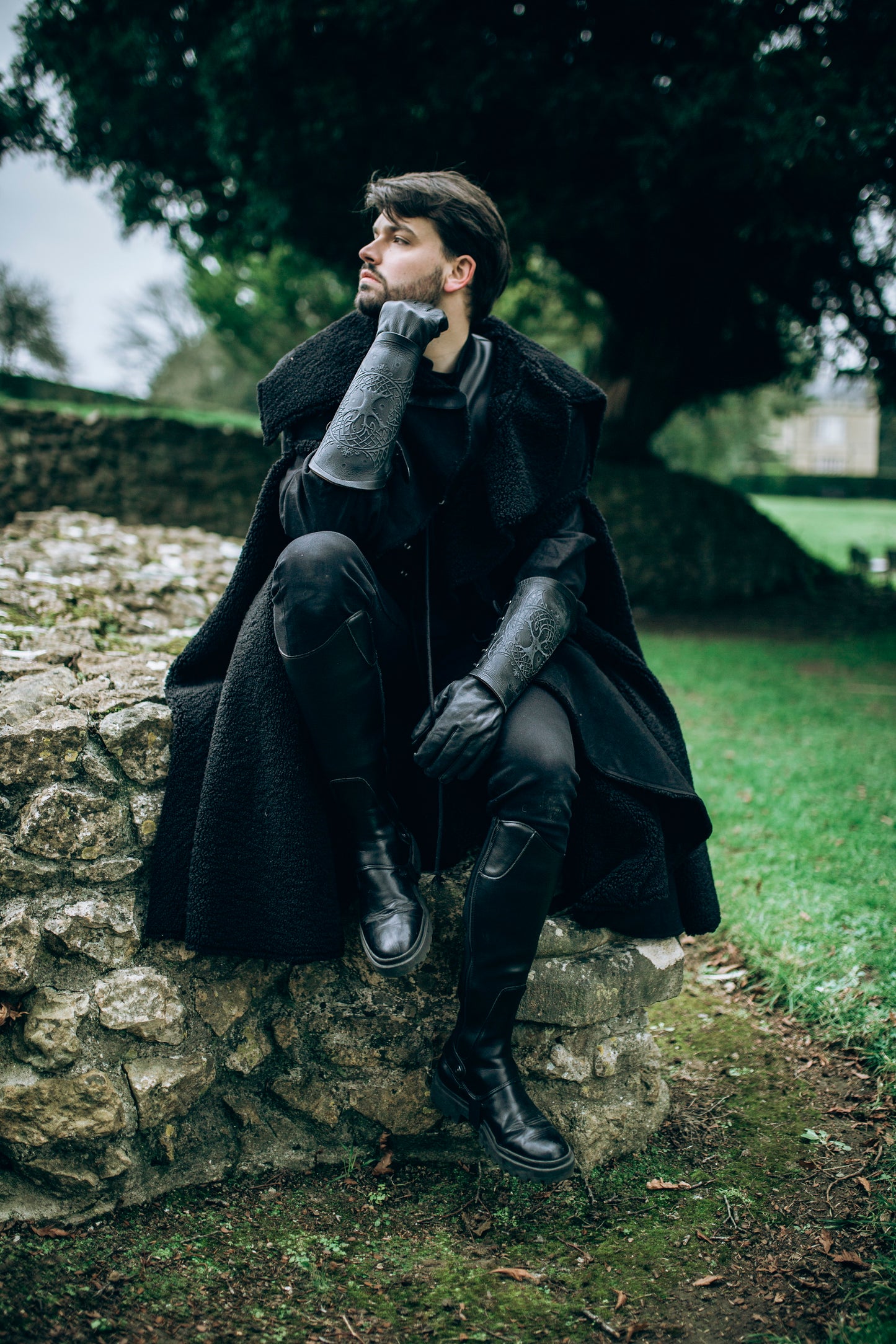 Fantasy adventurer wearing black leather and cloak sits on stone ruins, gazing into the distance. His arms feature long black leather bracers with Yggdrasil engraving, inspired by Norse mythology. This piece suits witcher armor, viking costume woman, or dark rogue cosplay. Perfect for LARP, ren faire, or medieval outfits, the bracers enhance any warrior or ranger look. The scenery evokes solitude and deep roleplay immersion in outdoor events.