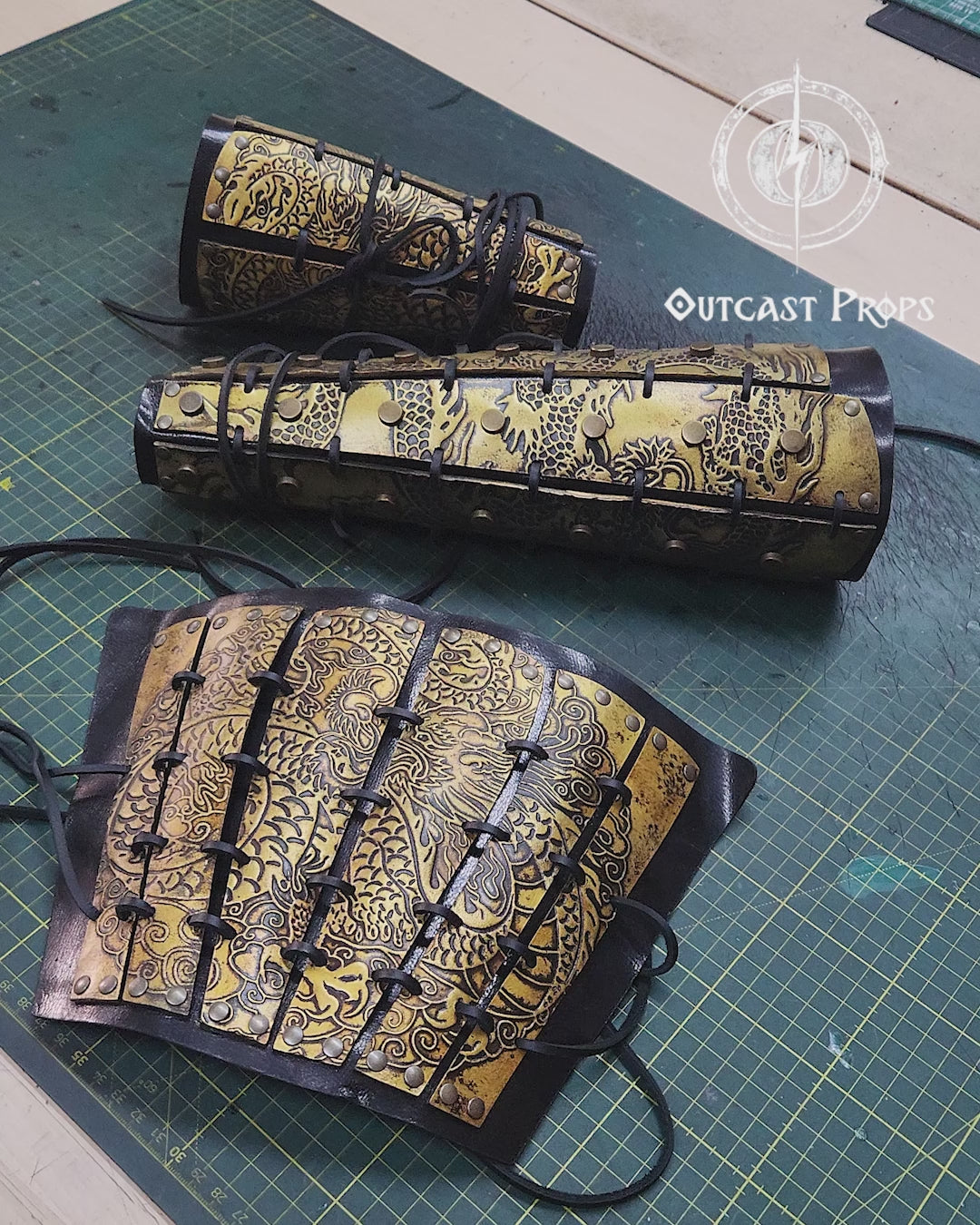 Golden engraved samurai armor bracers resting on a table with fantasy props like potions and maps. Designed for samurai larp costume and cosplay warriors. Black lacing and ornate dragon motifs give a powerful Eastern feel. Durable and handcrafted with vegetable-tanned leather and water-based dyes. Aged to resemble antique Japanese armor pieces. Great for L5A RPG or fantasy samurai characters in immersive games.