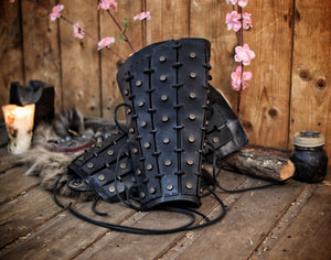 A pair of black leather samurai shin guards with studs and rivets, displayed against a rustic backdrop. Samurai Leather Greaves for LARP Cosplay or Renaissance Fair Studded Shin Armor Fantasy Warrior Leg Guards Handmade Medieval Costume Piece