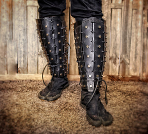 A pair of black leather samurai shin guards with studs and rivets, displayed against a rustic backdrop. Samurai Leather Greaves for LARP Cosplay or Renaissance Fair Studded Shin Armor Fantasy Warrior Leg Guards Handmade Medieval Costume Piece