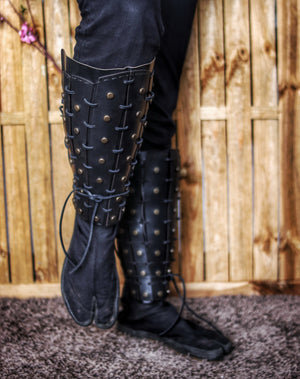 A pair of black leather samurai shin guards with studs and rivets, displayed against a rustic backdrop. Samurai Leather Greaves for LARP Cosplay or Renaissance Fair Studded Shin Armor Fantasy Warrior Leg Guards Handmade Medieval Costume Piece