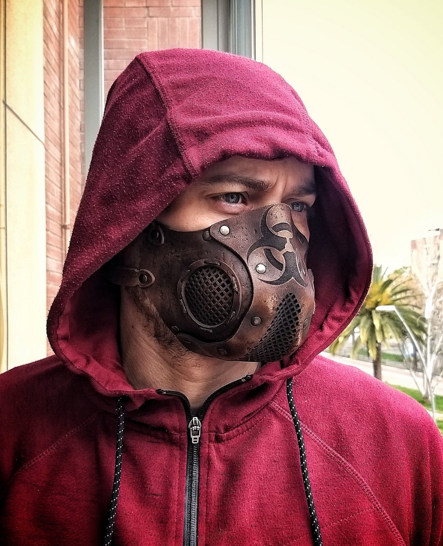 A person wearing a leather face mask with a hood, featuring a biohazard symbol and adjustable straps, in front of a background of foliage. Leather Face Mask with filter pocket Biohazard sign Post Apocalyptic mask for use with disposable face masks Paper towel or filter