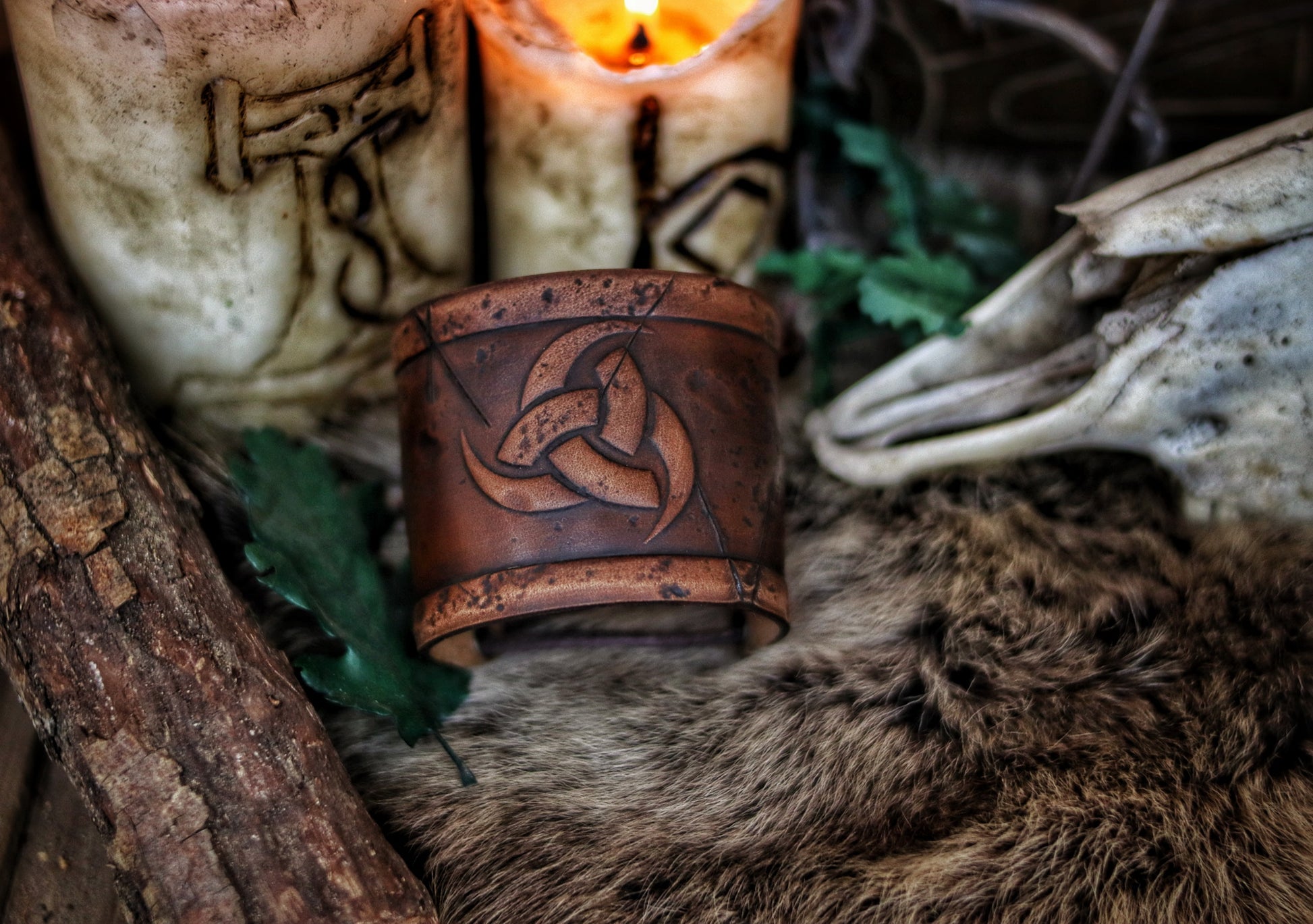Leather cuff bracelet with the Three Horns of Odin design, placed against a backdrop with candles and decorative foliage. Horns of Odin Leather Cuff Bracelet, Viking Bracer, leather embossed wristband, stamped viking pattern for LARP, cosplay and Fantasy fairs