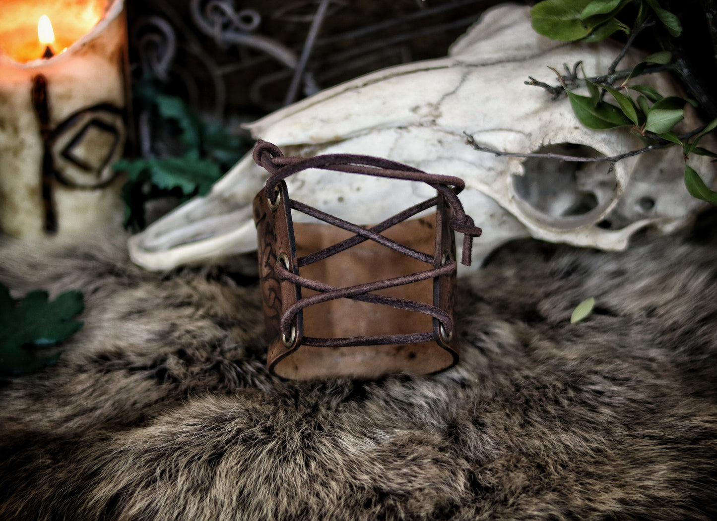Leather cuff bracelet with the Three Horns of Odin design, placed against a backdrop with candles and decorative foliage. Horns of Odin Leather Cuff Bracelet, Viking Bracer, leather embossed wristband, stamped viking pattern for LARP, cosplay and Fantasy fairs