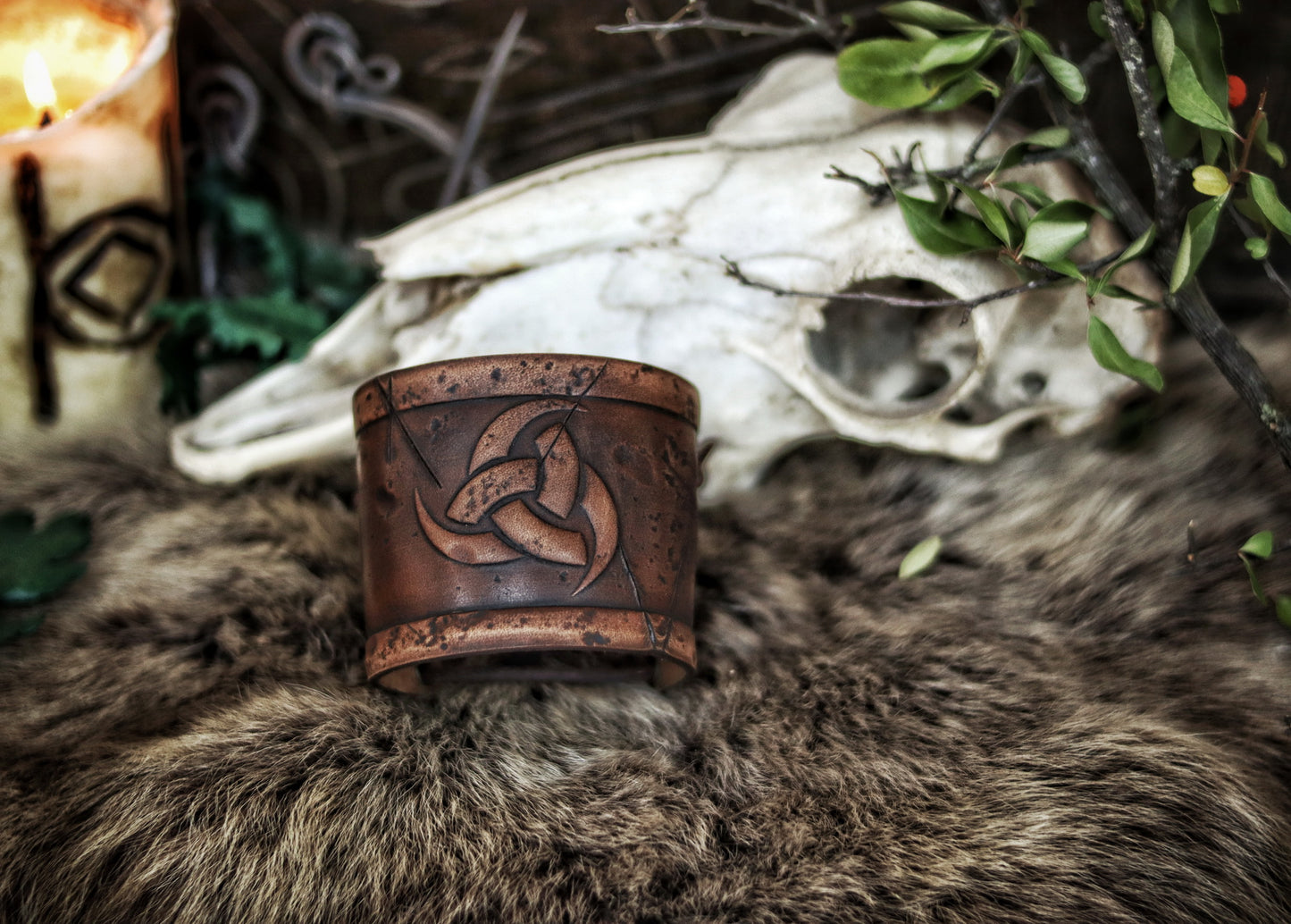 Leather cuff bracelet with the Three Horns of Odin design, placed against a backdrop with candles and decorative foliage. Horns of Odin Leather Cuff Bracelet, Viking Bracer, leather embossed wristband, stamped viking pattern for LARP, cosplay and Fantasy fairs