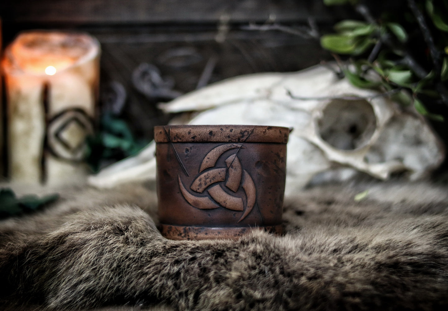 Leather cuff bracelet with the Three Horns of Odin design, placed against a backdrop with candles and decorative foliage. Horns of Odin Leather Cuff Bracelet, Viking Bracer, leather embossed wristband, stamped viking pattern for LARP, cosplay and Fantasy fairs