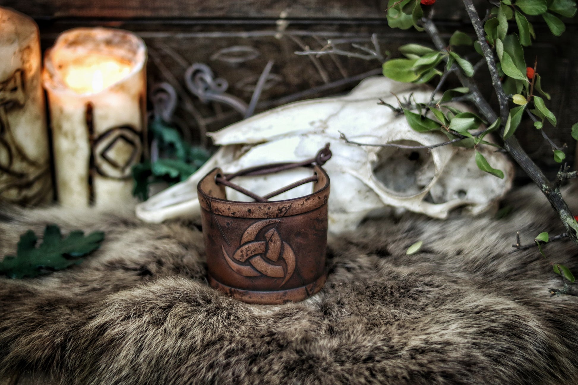 Leather cuff bracelet with the Three Horns of Odin design, placed against a backdrop with candles and decorative foliage. Horns of Odin Leather Cuff Bracelet, Viking Bracer, leather embossed wristband, stamped viking pattern for LARP, cosplay and Fantasy fairs