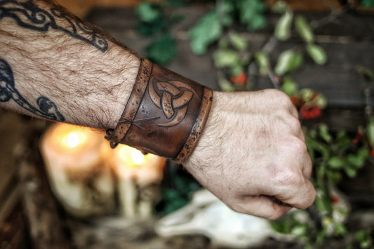 A person's forearm showcasing a leather cuff bracelet with the Three Horns of Odin design, placed against a backdrop with candles and decorative foliage. Horns of Odin Leather Cuff Bracelet, Viking Bracer, leather embossed wristband, stamped viking pattern for LARP, cosplay and Fantasy fairs