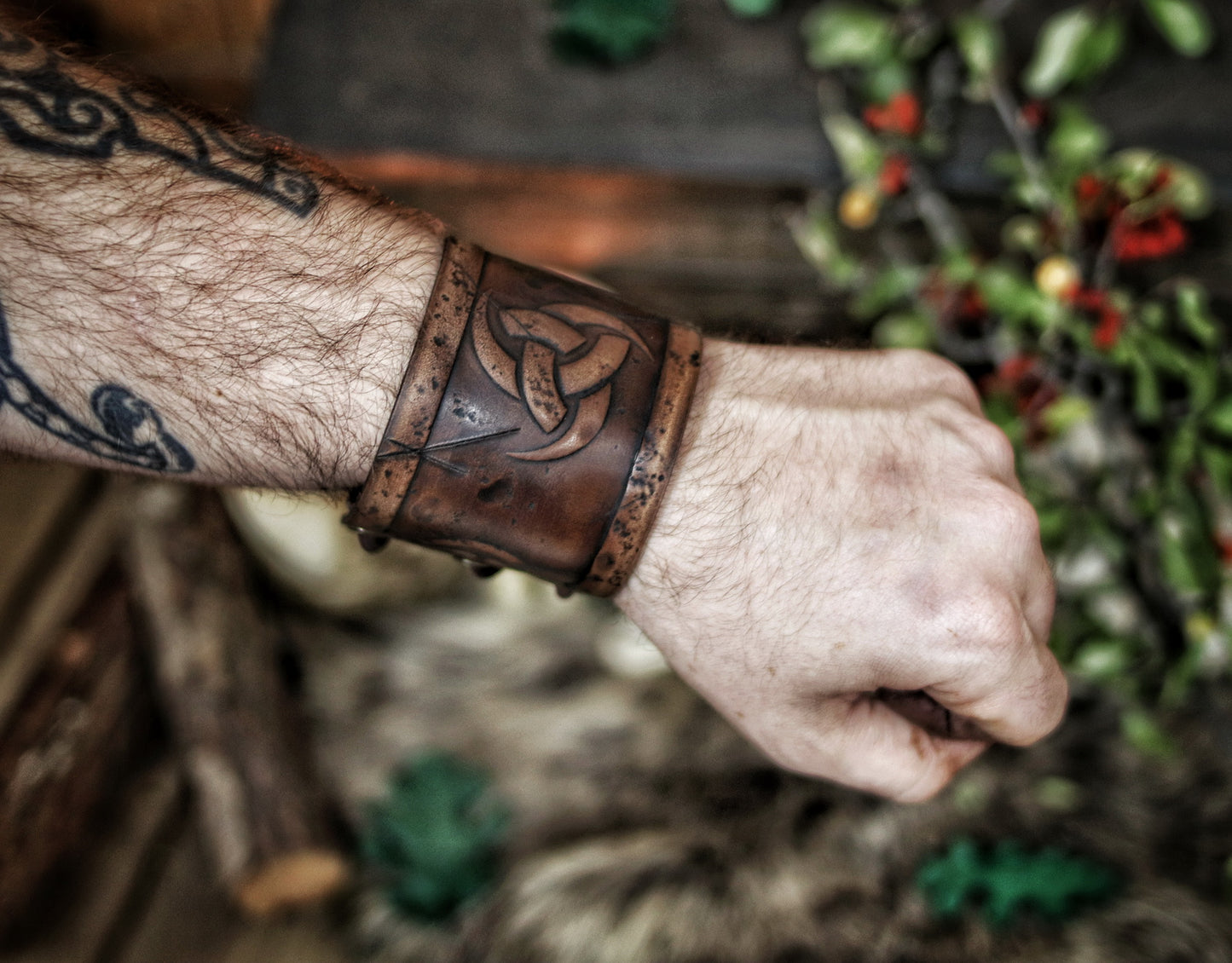 A person's forearm showcasing a leather cuff bracelet with the Three Horns of Odin design, placed against a backdrop with candles and decorative foliage. Horns of Odin Leather Cuff Bracelet, Viking Bracer, leather embossed wristband, stamped viking pattern for LARP, cosplay and Fantasy fairs