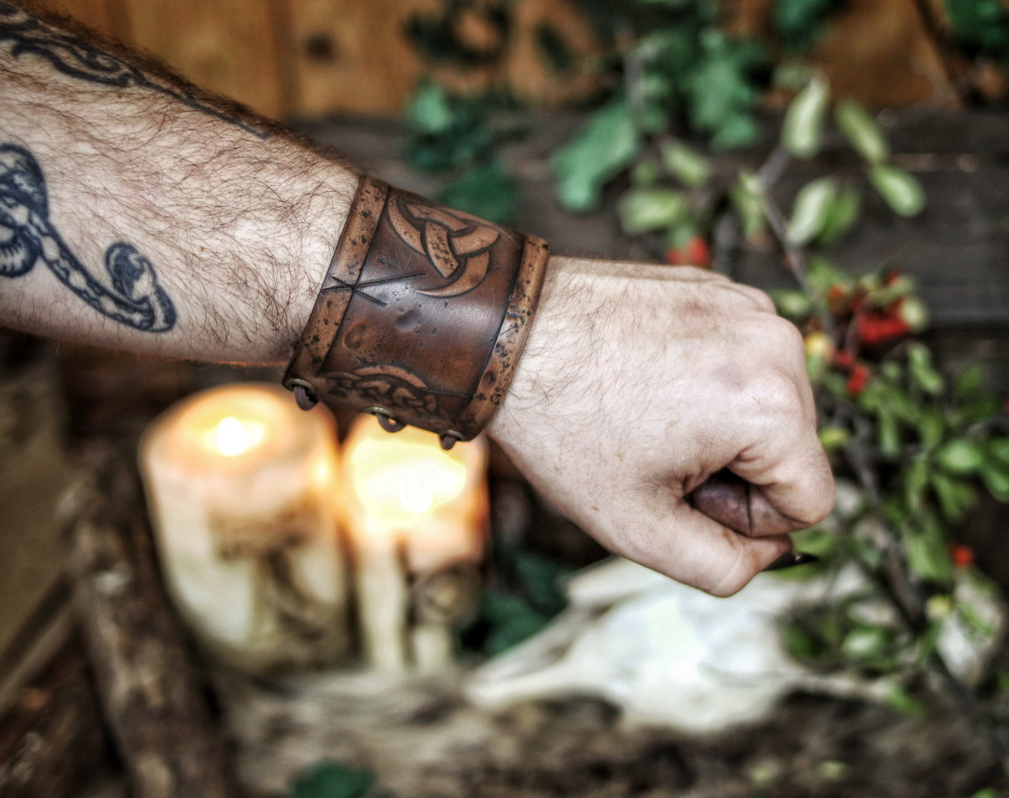 A person's forearm showcasing a leather cuff bracelet with the Three Horns of Odin design, placed against a backdrop with candles and decorative foliage. Horns of Odin Leather Cuff Bracelet, Viking Bracer, leather embossed wristband, stamped viking pattern for LARP, cosplay and Fantasy fairs