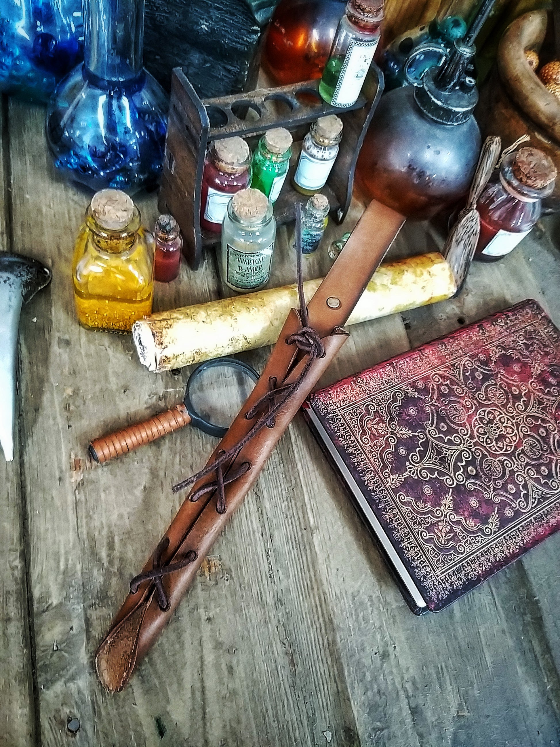 A close-up image of a brown wand holster with a leather strap and lace detail, displayed on an open book with a decorative background. Magic Wand Leather Holster Laced & Aged, perfect sheath for wizzards to hang from your belt, witch wand belt seath cosplay LARP