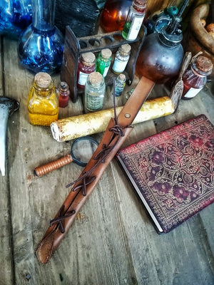 A close-up image of a brown wand holster with a leather strap and lace detail, displayed on an open book with a decorative background. Magic Wand Leather Holster Laced & Aged, perfect sheath for wizzards to hang from your belt, witch wand belt seath cosplay LARP